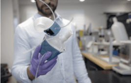 A 3d printed heart in the hands of a researcher. Half is blue and half is white. Clear tubes are stemming from the model to represent arteries.