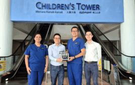 Four individuals stand smiling in front of the entrance to the Children's Tower at KK Women’s and Children’s Hospital. The person second from the left holds a small black device labeled BLIPI, designed for rapid point-of-care diagnostics. Two of the individuals wear blue medical scrubs, indicating their clinical roles.
