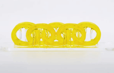 Yellow dual‑material test part, 3D‑printed by UT Austin’s two‑light system to demonstrate seamless soft‑and‑rigid sections.